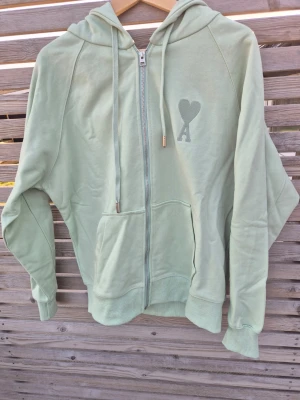 AMI Paris Hoodie - Snygg ljusgrön/turkos hoodie från Ami Paris med broderad AMI-logga på bröstet. Tillverkad i mjukt bomullstyg och har en klassisk huva. Aldrig använd/tvättad då jag tycker den är lite för stor.