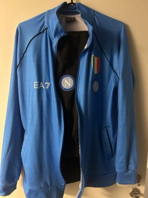 Napoli träningsdress från EA7 - Säljer en blå och svart träningsdress från EA7 x Napoli. Jackan har dragkedja, klubbmärke, EA7-logga och italienska mästerskapsmärket på bröstet. Byxorna är svarta med blå sidoremsor och Napoli-logga. Perfekt för fotbollsträning eller chill.