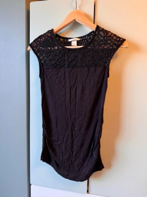 Svart ärmlös topp med spetsdetalj gravid - Gravid topp. Snygg svart ärmlös topp med spets upptill och rund halsringning. Toppen har en rak passform och är tillverkad i mjukt tyg med dekorativ spets över axlar och bröst. Det är XS men passade br mig som brukar ha S
