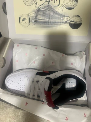 Nike Air Jordan 1 Low Vit/Röd/Svart - Snygga Nike Air Jordan 1 Low sneakers i vitt läder med svarta och röda detaljer. Klassisk perforerad tå, svart swoosh och röd Jumpman-logga på plösen. Yttersulan är svart och vit med tydlig Jumpman-logo. Perfekt för dig som gillar streetwear och ikonisk design.