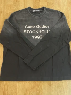Svart långärmad topp Acne Studios - Jag har kvitto på denna svarta långärmad topp från Acne Studios med vitt tryck på bröstet där det står Acne Studios STOCKHOLM 1996. Tillverkad i mjuk bomull och har en avslappnad passform. Perfekt för dig som gillar minimalistisk och trendig stil.