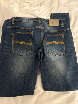 Blå jeans från Nudie Jeans - Säljer ett par klassiska blå jeans från Nudie Jeans med snygga slitningar och kontrastsömmar. Jeansen har raka ben, fem fickor och ikoniska broderade bakfickor med gul söm. Perfekta för en avslappnad och trendig stil.