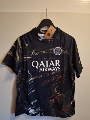 PSG x Jordan svart 2026 matchtröja - Snygg svart PSG matchtröja från Jordan med marmorerat mönster i guld och rött. Tröjan har korta ärmar, rund hals och tryckta loggor för PSG, Jordan och Qatar Airways. Perfekt för fotboll eller streetwear.