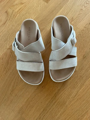  Nya Beige sandaler från Sweeks - Snygga beige sandaler från Sweeks med breda korslagda remmar i mocka och justerbart spänne på sidan. Sköna att bära med mjuk innersula och öppen tå. Perfekta för en avslappnad och trendig look.