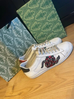 Gucci vita sneakers med orm-motiv - Snygga vita sneakers från Gucci med ikoniskt broderat orm-motiv i rött och svart på sidan. Skorna har vita snören, röd och grön detalj på hälen samt klassisk låg modell. Tillverkade i läder med gummisula. Perfekta för dig som vill sticka ut med lyxig streetstyle.