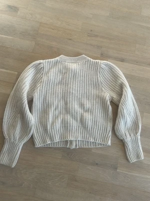 Beige stickad kofta från Vila - Supersöt beige stickad kofta från Vila med volangdetaljer framtill och stora knappar. Koftan har ballongärmar och ribbade muddar vid ärmslut och nederkant. Perfekt för lager-på-lager och en mysig look.