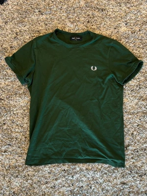 Mörkgrön t-shirt från Fred Perry - Snygg mörkgrön t-shirt från Fred Perry med klassisk logga broderad på bröstet. T-shirten har rund halsringning och korta ärmar. Perfekt för en stilren och avslappnad look.