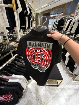 Svart Balmain Piké med tryck  - Svart Piké från Balmain med stort tryck och logga på ryggen i rött, vitt och grått. Framsidan har en liten Balmain-logga på bröstet. Klassisk passform med krage och korta ärmar. Endast använd ett fåtal gånger! Köpt av mig 2023 i Balmains butik i Dubai Mall där bilderna är tagna. Nypris 5 880 SEK då (1 960 AED). 
