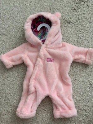 Rosa overall med björnöron för dockor - Supermjuk rosa overall i plysch med huva och små björnöron. Perfekt för dockor, overallen har ett rutigt innerfoder i huvan och stängs framtill. Söt detalj med namnlapp med Lillan på.