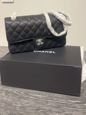 Väska i svart  - Ikonisk svart  Classic Flap Bag i quiltat läder med silverfärgad CC-låsning och kedjerem. Klassisk rektangulär form och elegant design, perfekt för dig som vill ha en tidlös och lyxig handväska. Levereras med originalbox.