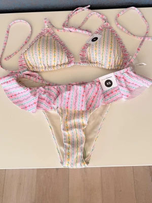 Blommig bikini med volang från Bikbok - Supersöt bikini från Bikbok, överdel storlek S och underdel storlek XS med blommigt mönster i rosa, gul och grönt. Överdelen har inlägg som går att ta ut och med knytband i nacke och rygg, och underdelen har volangdetalj runt hela midjan. Köptes förra sommaren men kom aldrig till användning, alla lappar och plaskydd är kvar!! Nypris 400kr