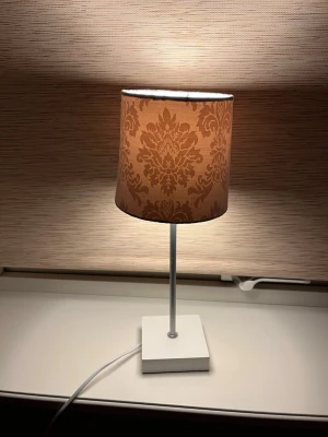 Bordslampa med mönstrad skärm - Snygg bordslampa med beige mönstrad lampskärm och vit fyrkantig fot. Lampan har en smal metallstång och tygsladd. Perfekt som mysbelysning i sovrum eller vardagsrum.köpt för 350