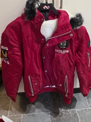 Röd pufferjacka från Leduntino  - Röd pufferjacka med broderade detaljer och patchar från Leduntino. Jackan har dragkedja, knappar, två framfickor med dragkedja och fuskpäls på huvan. Fodrad med vit teddy på insidan för extra värme. Snygga emblem på bröstet och ärmen.