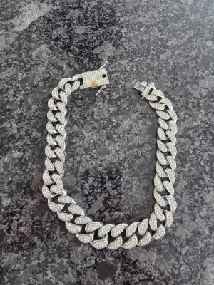 Blingigt silverfärgat cuban chain halsband - Säljer ett riktigt blingigt cuban chain halsband i silverfärg med massor av gnistrande stenar. Halsbandet har breda länkar och ett rejält lås täckt av små stenar för extra lyxig känsla. Perfekt för dig som vill sticka ut och addera lite extra shine till din outfit.
