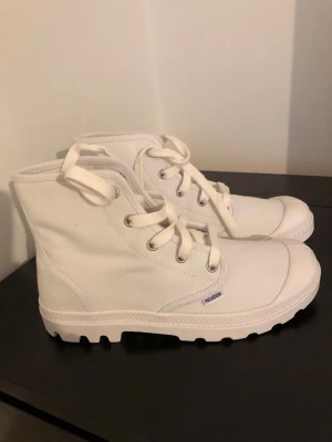 Vita canvas kängor från Palladium - Snygga vita kängor från Palladium i canvasmaterial med grov sula och klassisk snörning. Perfekta för dig som gillar en clean och stilren look. Märkesdetalj på sidan och robust design som passar till många outfits.