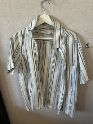 Randig kortärmad skjorta från H&M - Snygg kortärmad skjorta från H&M i vitt med svarta och gråa ränder. Skjortan har klassisk krage, knäppning framtill och bröstficka. Tillverkad i ett lätt och luftigt linnematerial, perfekt för varma dagar.