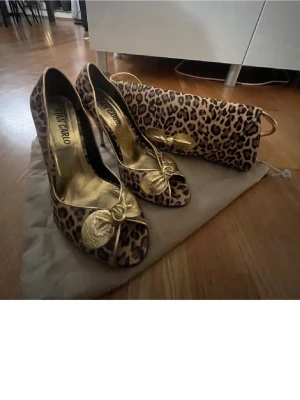 Leopardmönstrade pumps från San Carlo - Snygga pumps med leopardmönster och guldfärgade detaljer från San Carlo. Skorna har öppen tå och en dekorativ rosett framtill. Insidan är guldfärgad och klacken är hög. Perfekt för dig som gillar att sticka ut med djurmönster.