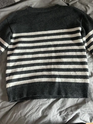 Mörkgrå randig stickad tröja - Mysig stickad tröja i grått och vitt med horisontella ränder över hela kroppen och ärmarna. Tröjan har rund halsringning och ribbade muddar vid ärmslut och nederkant.