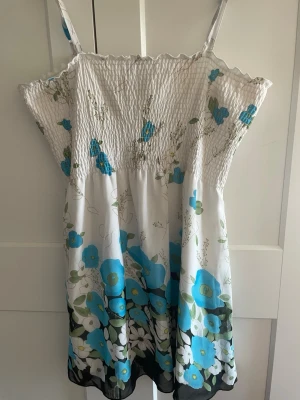 Blommig sommarklänning från Milla - Söt kort klänning från Milla med vita och blå blommor, smock upptill och tunna axelband. Perfekt för varma dagar med sitt lätta material och fina blommönster. Klänningen har en avslappnad passform och ärmlös design.