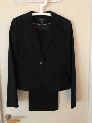 Svart kavaj från H&M, storlek 36 - Stilren svart kavaj från H&M i klassisk modell med en knapp framtill och smala slag. Kavajen har diskreta fickor och är tillverkad i ett mjukt syntetmaterial. Perfekt för en clean och snygg look.