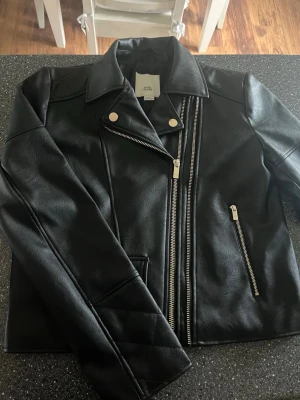 Svart skinnjacka från river island - Snygg svart skinnjacka från river island med klassisk bikerstil, sneddragna dragkedjor och silvriga detaljer. Jackan har två fickor med dragkedja och en dekorativ ficka framtill. Perfekt för dig som vill ha en cool och tidlös look. Använd en gång och nu är för liten!