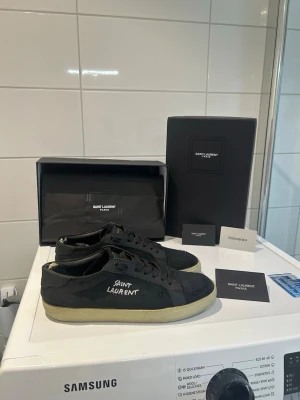 Svarta Saint Laurent sneakers - Svarta sneakers från Saint Laurent med vit logga på sidan och svarta skosnören. Ovandel i canvas och detaljer i läder, med slitna kanter för en cool vintage-look. Klassisk låg modell med vit sula. Kommer med originalkartong och tillbehör. Skorna är i mycket bra skick bara att insidan är lite slitet men inget som märks av 
