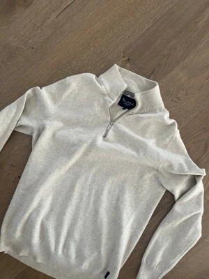 Beige halvzip-tröja från Bondelid - Snygg beige stickad tröja från Bondelid med halv dragkedja. Tröjan har diskret broderad logga på bröstet och ribbade muddar vid ärmslut och nederkant. Perfekt för en stilren och avslappnad look.