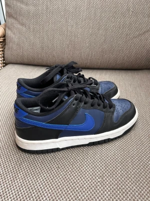 Nike Dunk Low svart/blå sneakers - Säljer ett par Nike Dunk Low sneakers i svart och blått med vit sula. Skorna har klassisk låg siluett, svart snörning och perforerad tåbox för extra ventilation. Swoosh-loggan på sidan i blått och ovandel i läder ger en clean och sportig look.