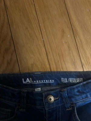Mörkblå regular fit jeans LAB Industries - Snygga mörkblå jeans från LAB Industries i modellen Felix Regular Fit. Klassisk femficksdesign med raka ben och knappgylf. Perfekta för en avslappnad och stilren look. Tillverkade i slitstarkt denimtyg.
