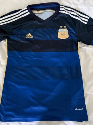 Argentina fotbollströja Adidas blå - Snygg blå Argentina fotbollströja från Adidas med guldiga detaljer och två stjärnor ovanför klubbmärket. Tröjan har vita ränder på axlarna, V-ringning och är tillverkad i lätt och ventilerande climacool-material. Perfekt för fotbollsträning eller match.