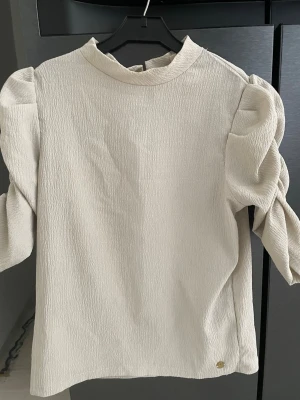 Pernilla Wahlgren Beige blus med puffärmar - Pernilla Wahlgren Stilren beige blus med puffiga långärmar och lätt rynkad struktur. Hög halsringning med liten volangkant och knapp i nacken. Tillverkad i ett krinklat tyg som ger en trendig look. Perfekt för dig som gillar en chic och modern stil.