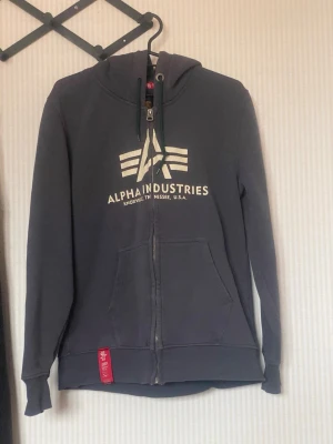 Mörkgrå hoodie från Alpha Industries - Snygg mörkgrå hoodie med dragkedja från Alpha Industries. Stor vit logga och text på bröstet, två fickor framtill och klassisk huva med snörning. Röd detalj vid nederkanten. Perfekt för en avslappnad streetstyle-look.
