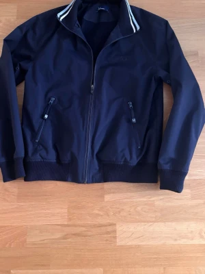Mörkblå vindjacka med dragkedja -  Tjena säljer en riktigt fet Fred Perry mörkblå vindjacka. Den har dragkedja, två snedställda fickor med dragkedja och ribbade muddar vid ärmar och midja. Kragen har vita ränder för en fet look. Perfekt för blåsiga dagar. Riktigt fet när lufter blir kallare på kvällarna