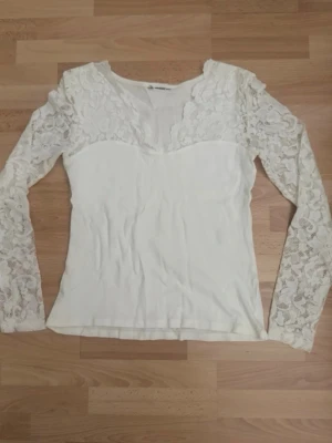 Vit spetsblus med lång ärm - Snygg vit blus med långa ärmar i spets och v-ringad halsringning. Överdelen och ärmarna har ett vackert spetsmönster som ger en romantisk känsla. Perfekt att styla med jeans eller kjol för en trendig look. Lapparna bortklippta. Bröst 80-86cm.