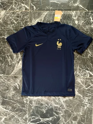 Blå Nike Frankrike matchtröja - Snygg mörkblå matchtröja från Nike med Frankrikes landslagsmärke och två stjärnor på bröstet. Tröjan har korta ärmar, rund hals och är tillverkad i ett lätt, funktionellt material som passar perfekt till träning eller match.