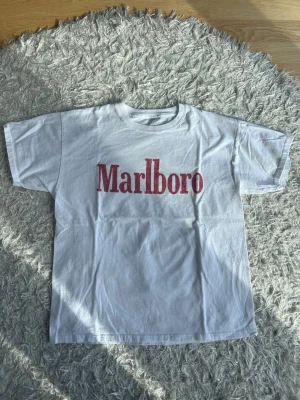 Vintage Marlboro Tshirt - Vit t-shirt från Marlboro med klassisk röd logga tryckt på bröstet. T-shirten har rund hals och korta ärmar, perfekt för en avslappnad och cool stil. Materialet känns som bomull och passformen är normal. Sitter som en medium
