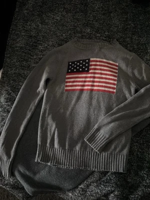 Grå stickad tröja med USA-flagga - Snygg grå stickad tröja med en stor amerikansk flagga på bröstet. Tröjan har rund halsringning, långa ärmar och ribbstickade muddar vid ärmslut och nederkant. Perfekt för dig som gillar streetstyle och vill sticka ut.