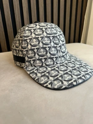 Mönstrad keps från FRGMO - Säljer en snygg keps från Salvatore Ferragamo med grått och vitt all-over-mönster och svart läderdetalj bak. Kepsen har böjd skärm och justerbart spänne bak. Tillverkad i canvasmaterial som ger en exklusiv känsla och passar perfekt till streetwear eller casual outfits.