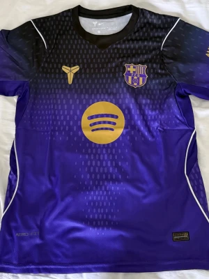 Lila FC Barcelona matchtröja - Säljer en lila och svart FC Barcelona matchtröja med guldiga detaljer, klubbmärke och sponsorloggor. Tröjan har korta ärmar, rund hals och är tillverkad i ett lätt, ventilerande funktionsmaterial. Perfekt för fotbollsträning eller att bära som supporter.
