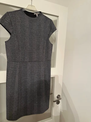Grå kortärmad klänning med mönster - Snygg grå klänning med korta ärmar och diskret fiskbensmönster. Modellen är figurnära med rund halsringning och dragkedja i ryggen. Perfekt för dig som gillar stilrena och klassiska plagg. Aldrig använd dock tvättad en gång:)