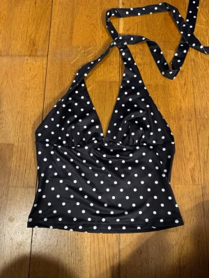 Svart baddräkt med vita prickar S - Snygg svart baddräkt från RIDER med vita prickar och halterneck-knytning. Klassisk retrostil med vadderade kupor och mjukt, stretchigt material som sitter bekvämt. Perfekt för strand eller poolhäng.