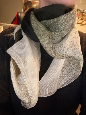Tunn halsduk i melerad färger  - Snygg halsduk i tunnt, transparent material med grönt, svart och vit melerad färg. Den har en lätt skimrande yta. Finns ett litet hål men inget som syns när man bär den.