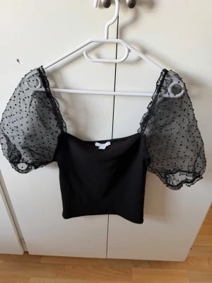 Svart topp med puffärmar från Amisu - Trendig svart topp från Amisu med transparenta puffärmar i mesh och små prickar. Toppen har en figurnära passform och fyrkantig urringning. Perfekt för dig som vill sticka ut med en unik detalj på ärmarna.