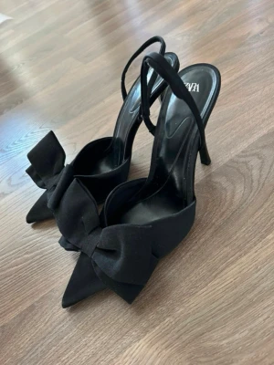 Svarta slingbacks med rosett från Zara - Stilrena slingbacks i fint skick från Zara med spetsig tå och rosett framtill. Klackhöjd ca 10cm.