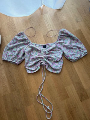 Blommig croppad topp från Gina Tricot - Superfin blommig topp från Gina Tricot med puffiga korta ärmar och snörning framtill. Toppen är croppad och har ett färgglatt mönster i rosa, grön och gul på ljus botten. Perfekt för dig som gillar romantisk och trendig stil.
