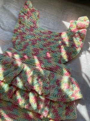 Handvirkad babyromper 50–56 100% bomull - Handvirkad babyromper i mjukt bomullsgarn. Pastellfärgad i rosa, mintgrönt och ljusgult med volanger och knappar nedtill. Storlek ca 50–56, nyfödd till cirka 2 månader. Mått: bredd midja ca 19 cm platt, längd ca 34 cm. Eftersom plagget är handgjort rekommenderas att jämföra måtten.