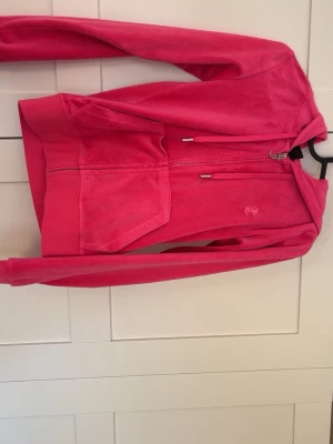 Juicy Couture rosa hoodie - Juicy Couture hoodie i en stark rosa färg, tillverkad i mjuk velour. Klassisk modell med dragkedja, huva och känguruficka framtill. Perfekt för en chill och trendig look. Orginalpris 1300kr.