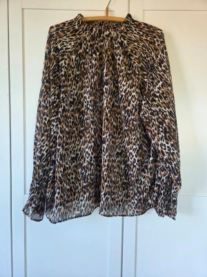 Leopardmönstrad blus med volangärm, XL - Snygg blus med leopardmönster i brunt, svart och beige. Blusen har långa ärmar med lätt volang vid ärmslut och en lös, luftig passform. Perfekt för dig som gillar djurmönster och vill sticka ut lite extra. Materialet känns tunt och flowy.