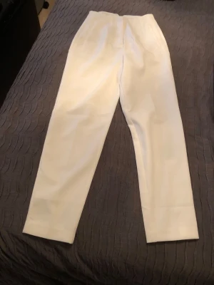 Vita kostymbyxor med rak passform - Snygga vita kostymbyxor med rak passform och pressveck. Byxorna har diskreta fickor bak och är tillverkade i ett slätt tyg som ger en stilren look. Perfekta för dig som vill ha en clean och modern stil.