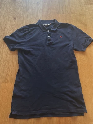 Mörkblå pikétröja från Abercrombie - Klassisk mörkblå pikétröja från H&M med korta ärmar, krage och knappar framtill. Liten röd logga broderad på bröstet. Perfekt till jeans eller shorts för en avslappnad stil. Tveka inte att lägga bud!